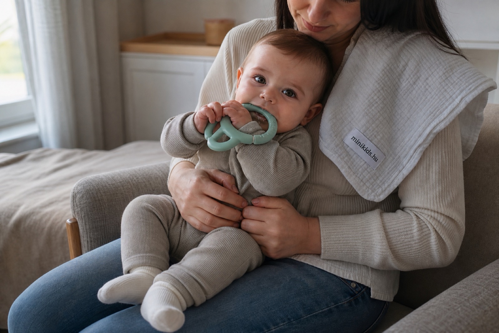 Anyuka ölében ülő, nyolc hónap körüli baba hűtött szilikon rágókát rág, két kezében szorongatja