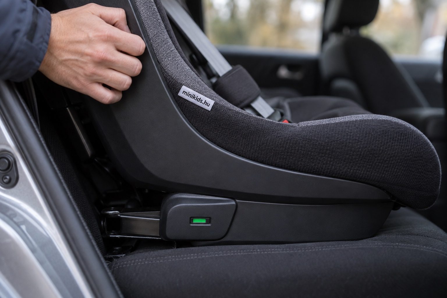 Közeli kép egy autóba szerelt gyerekülés oldaláról, ahol zöldre váltott az ISOFIX jelzője, mellette egy felnőtt keze fogja az ülés vázát