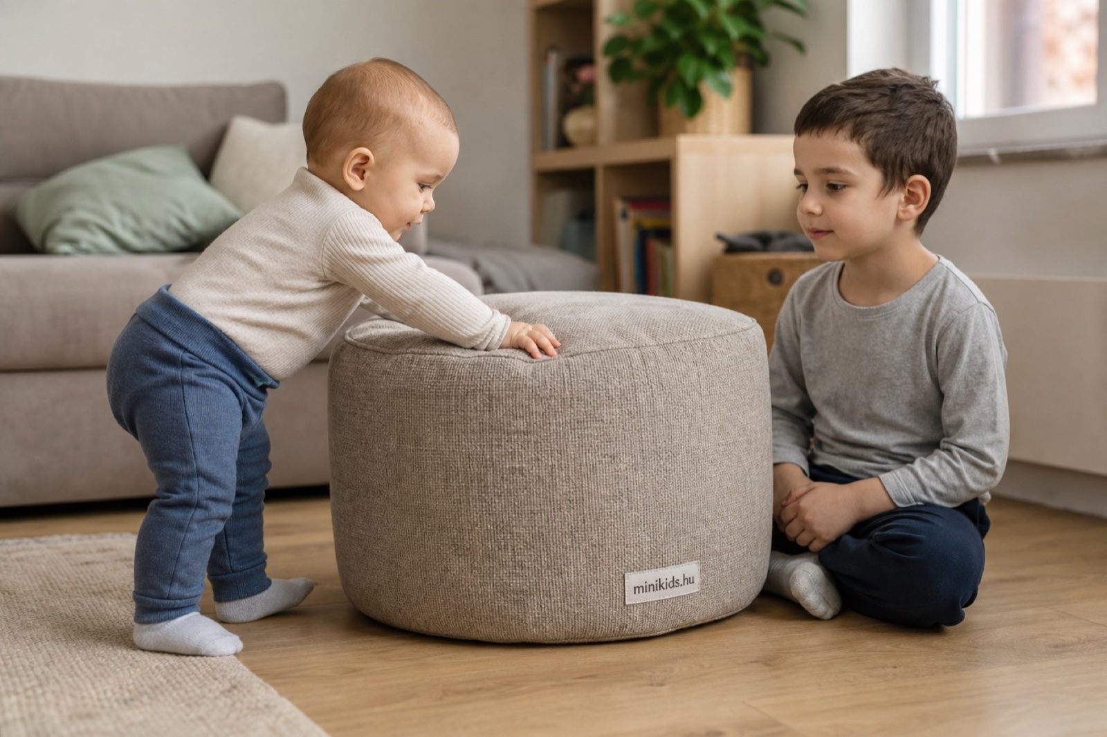 Zokniban álló baba egy stabil, puha pufra kapaszkodva húzza fel magát a nappaliban, mellette a padlón ülő testvére figyeli