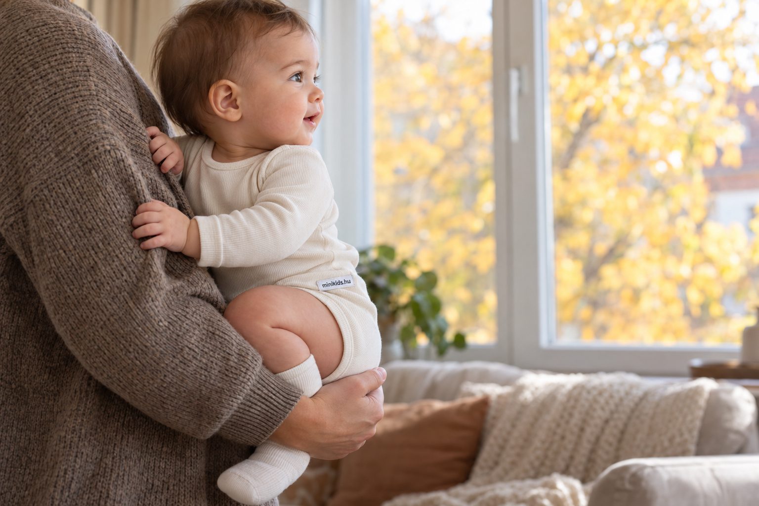 Szülő karján ülő baba oldalnézetből a nappali ablaka előtt, őszi délelőtti fényben, a baba mindkét keze a szülő pulóverén nyugszik